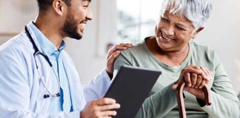 A doctor is holding a tablet computer, with one hand on the shoulder of an elderly patient. They are both smiling while looking at the test results.