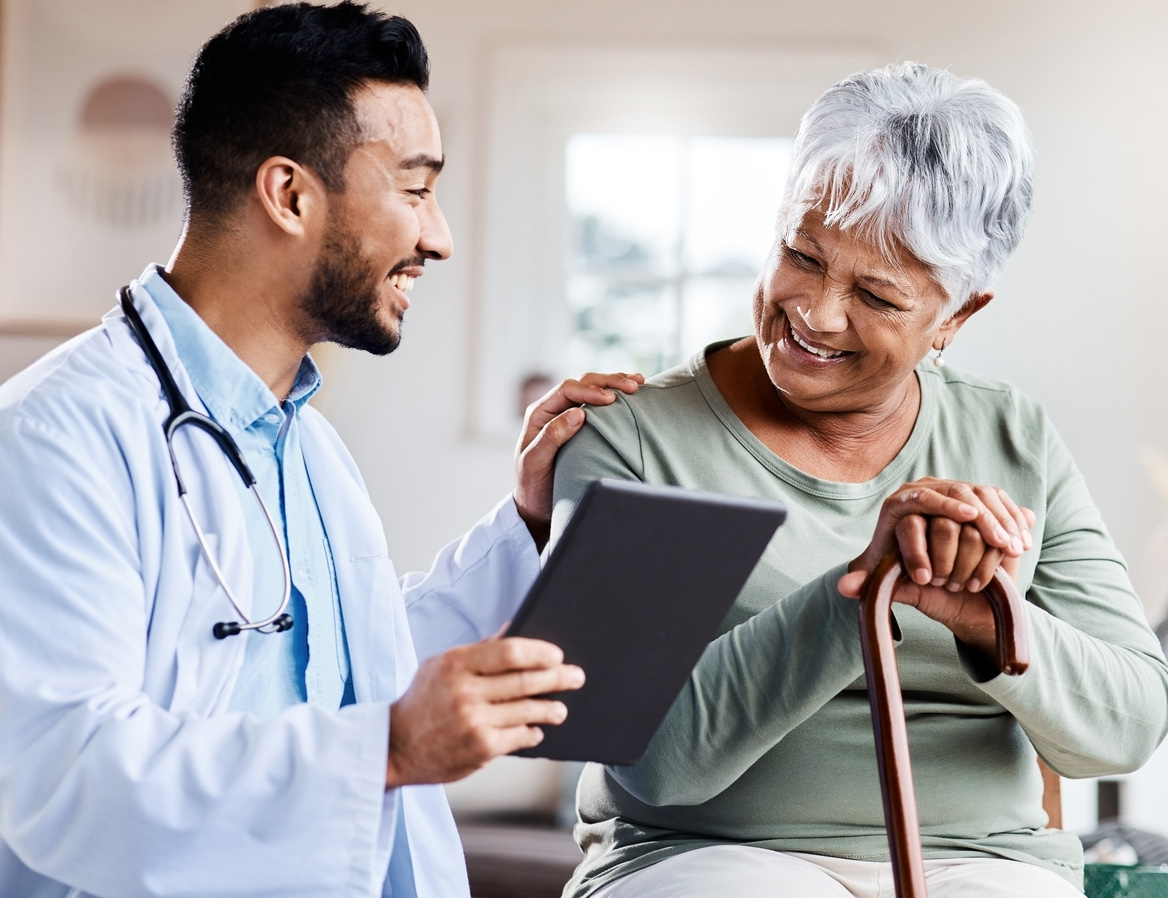 A doctor is holding a tablet computer, with one hand on the shoulder of an elderly patient. They are both smiling while looking at the test results.