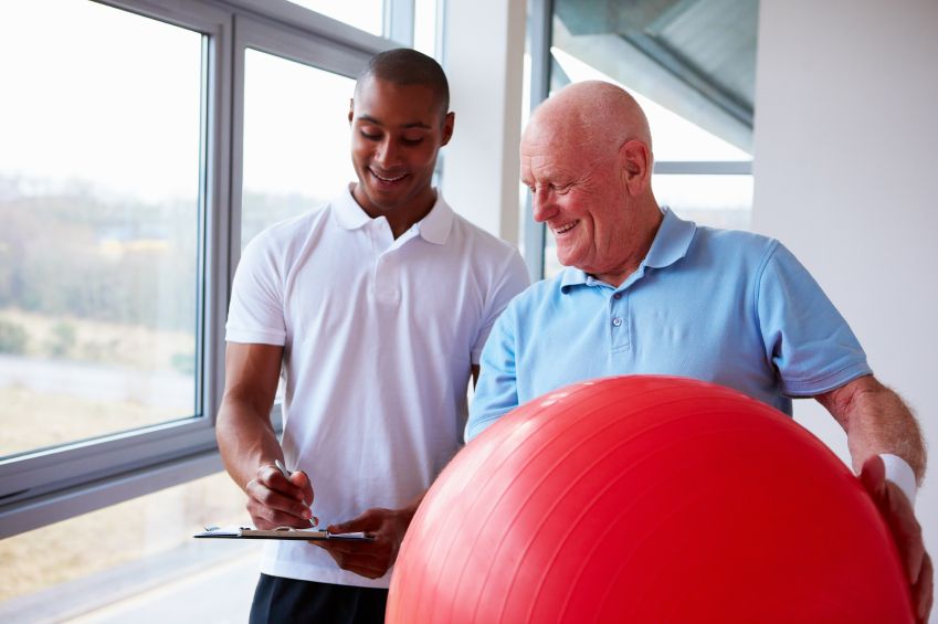 Rehab specialist looking at clipboard together with a client that is holding a large red fitness ball